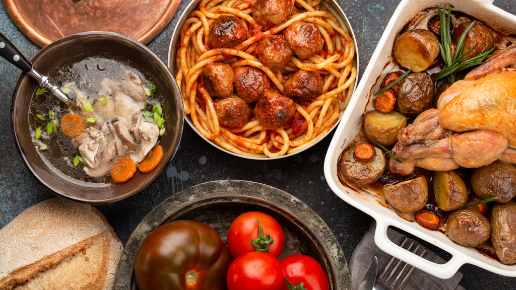 Overhead view of a hearty meal featuring spaghetti with meatballs, roasted chicken with potatoes, homemade soup, fresh tomatoes, and rustic bread.